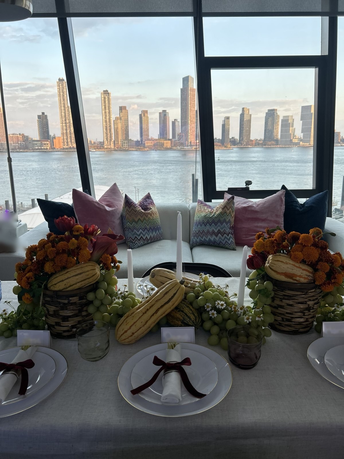 A harvest tablescape of French marigolds, tulips and delicata squash in woven baskets, green grapes and chamomile running down the table, white taper candles, and a river skyline through the windows